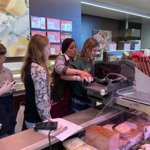 Schülerin schneidet Wurst in der Feinkost. Eine Mitarbeiterin hilft ihr. 