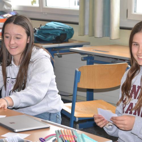 Zwei Schülerinnen lächeln in die Kamera, während sie eine Aufgabe erledigen. 