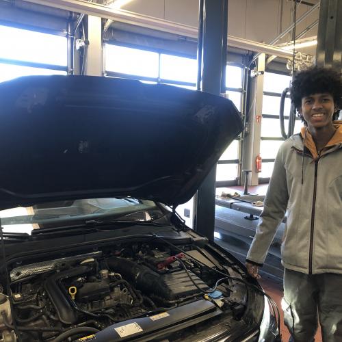 Ein Schüler steht vor einem Auto mit geöffneter Motorhaube.