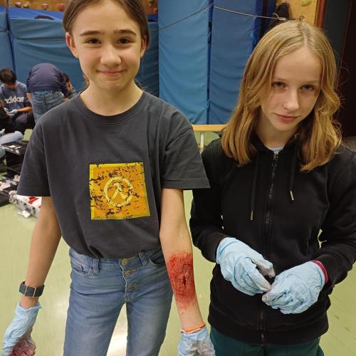Eine Schülerin bekam eine Wunde am Arm geschminkt. Eine Schülerin steht mit Gummihandschuhen daneben. Bereit sie zu "verarzten".