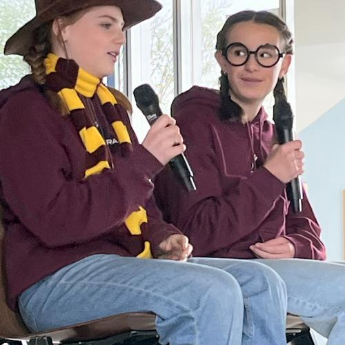 Zwei Schülerinnen sitzen auf der Bühne bei den Abschlusspräsentationen. Ihre Outfits erinnern an Harry Potter.