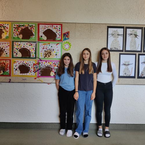 Drei Schülerinnen stehen vor einer mit von Kindern gezeichneten Bildern beklebten Wand.