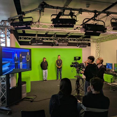 Zwei Schüler stehen vor einem Greenscreen und ein Schüler bedient die Kamera.