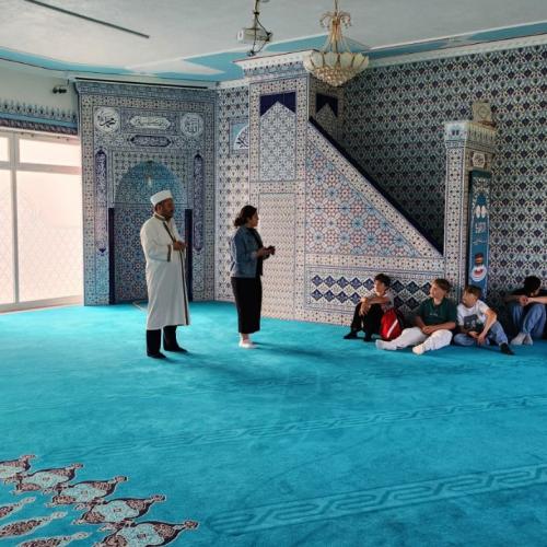 Besuch in der Moschee. Auf dem Bild ist der Imam mit einer Schülerin zu sehen. Rund herum sitzen Kindern im Kreis.