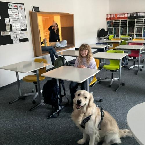 Bingo sitzt im Klassenzimmer und zwei Schülerinnen sind im Hintergrund zu sehen.