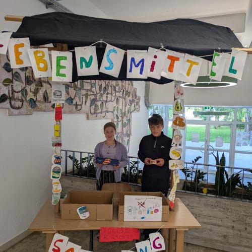 Zwei Schüler:innen an ihrem selbstgestalteten Stand für die Lebensmittelsammlung in der Schule.